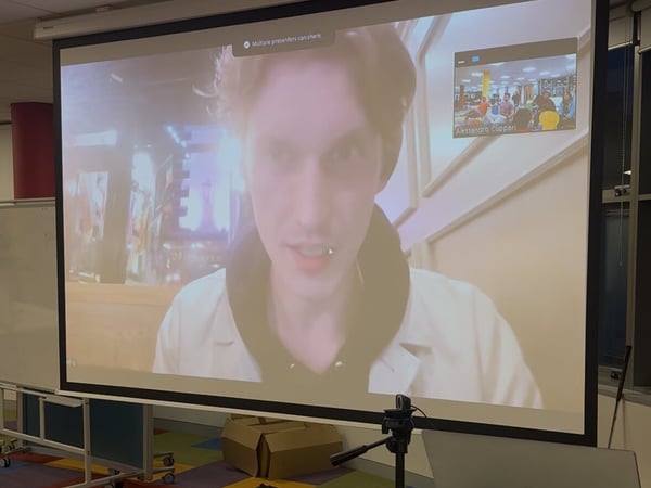 A photograph capturing a large projector screen displaying a video conference, featuring a close-up of a male speaker and a smaller inset window showing a group of people in a brightly colored room.