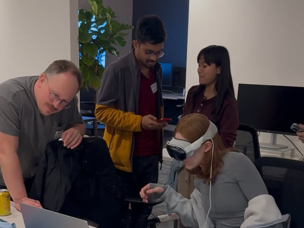 Four people are gathered in an indoor office setting, observing a woman seated at a desk who is wearing a white VR/AR headset and appearing to interact with a virtual environment using hand gestures.