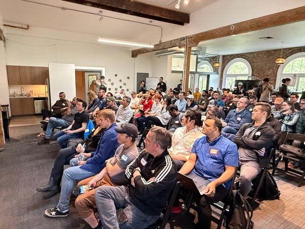 A man in a white t-shirt is giving a presentation to a large, seated audience in a modern office space with exposed brick walls and wooden beams.