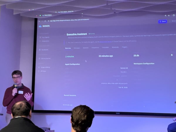 A man is giving a presentation in front of a large screen displaying a software dashboard for an AI 'Executive Assistant'.