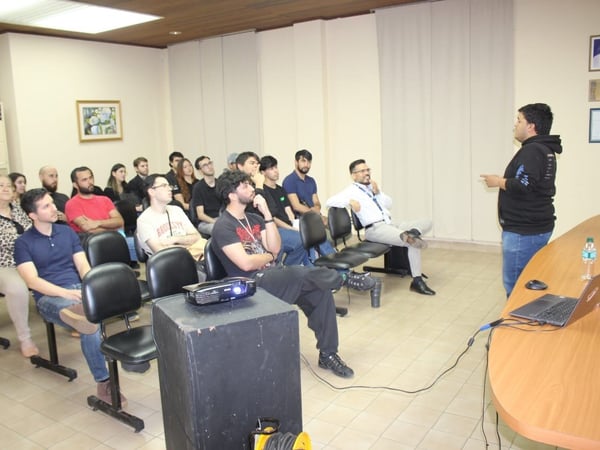 A candid photograph captures a group of approximately eighteen people seated in rows, attentively watching a standing presenter in an indoor conference or classroom setting.