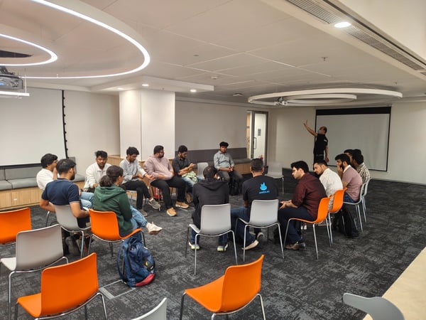 A group of approximately fifteen people are gathered in a modern office meeting room, seated in a circular arrangement of chairs for a discussion or training session, with one person standing and gesturing near a projection screen.
