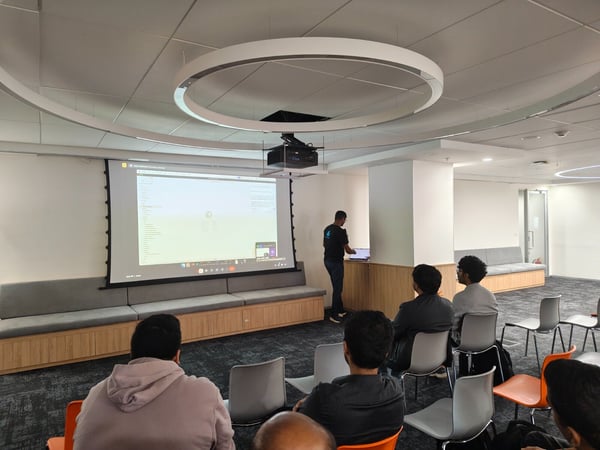 A group of attendees watches a technical presentation projected onto a large screen in a modern meeting room, while the presenter stands to the side operating a laptop.