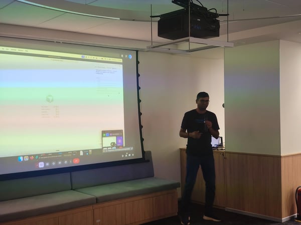 A photograph captures a man standing in front of a large, brightly lit projection screen, giving a presentation or talk in a modern indoor meeting space with built-in wooden bench seating.