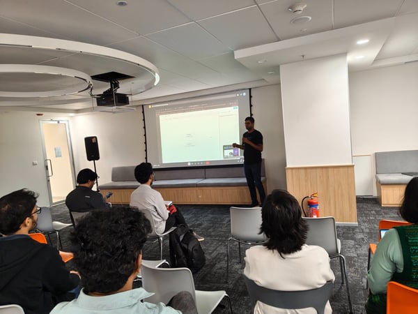A man is giving a presentation in a modern office or meeting room, standing next to a large projected screen while an audience is seated and watching.