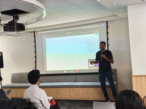 A man wearing glasses and a black shirt is giving a presentation in a modern office setting, standing in front of a large projection screen displaying a technical diagram, while an audience watches from the foreground.