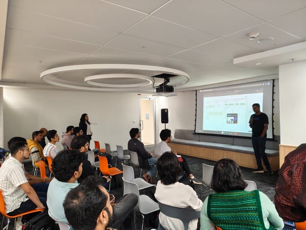 A photograph capturing an indoor presentation or meeting where a man is speaking to a seated audience in front of a large projection screen displaying a technical dashboard with 'No data' warnings.