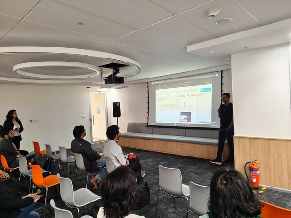 A photograph taken indoors shows a man giving a technical presentation using a large projected screen to a small audience seated in a modern office or conference room setting.