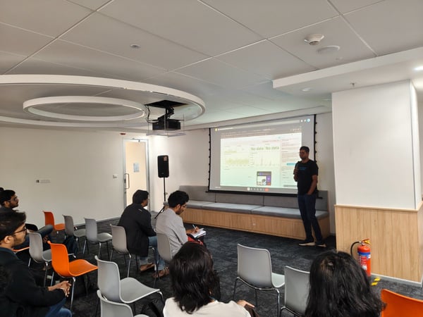 A photograph showing an indoor presentation event in a modern office space, where a speaker stands next to a large projected screen displaying a dashboard interface with the text 'No data No data' visible.
