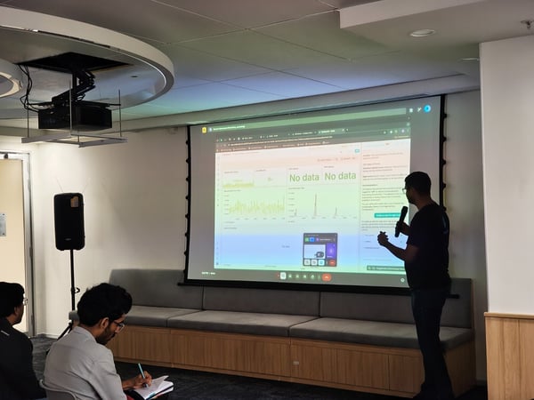 A photograph capturing an indoor technical presentation where a man, holding a microphone, stands next to a large projected screen displaying charts and data dashboards. Two attendees are visible in the foreground, seated and focused, with one taking notes.