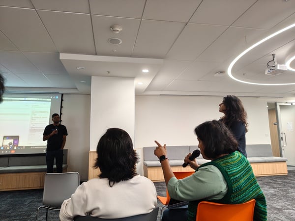 A candid photograph captures an indoor presentation or Q&A session where a man stands near a projection screen while an audience member in the foreground holds a microphone and gestures during a discussion.