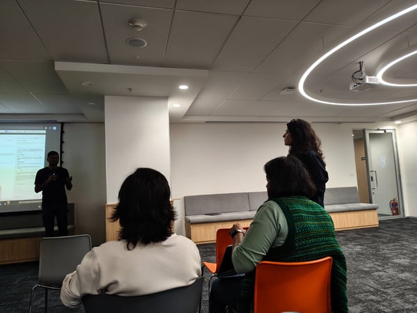 A candid photograph taken indoors during a technical presentation, showing a presenter silhouetted against a projection screen displaying code, while three audience members are visible in the foreground, mostly seen from behind.