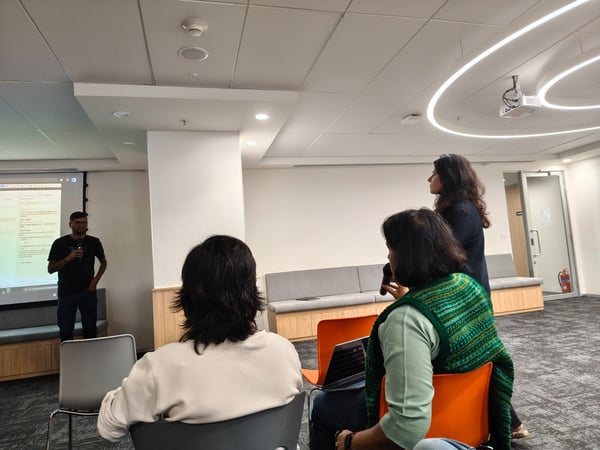 A candid photograph captures a presentation or discussion in a modern office meeting room, showing a man speaking near a projector screen while several attendees sit facing away from the camera.