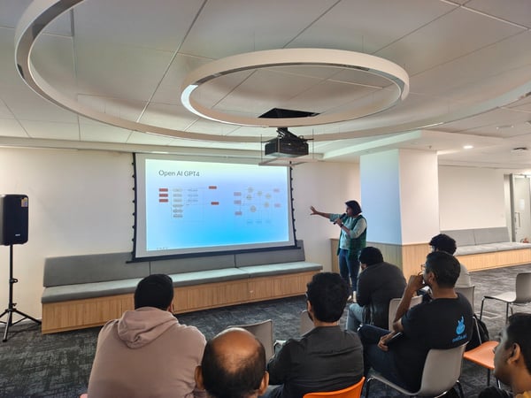 A presenter stands next to a large projection screen displaying a flow chart titled "Open AI GPT4," addressing a group of attendees seated in a modern, well-lit indoor space.