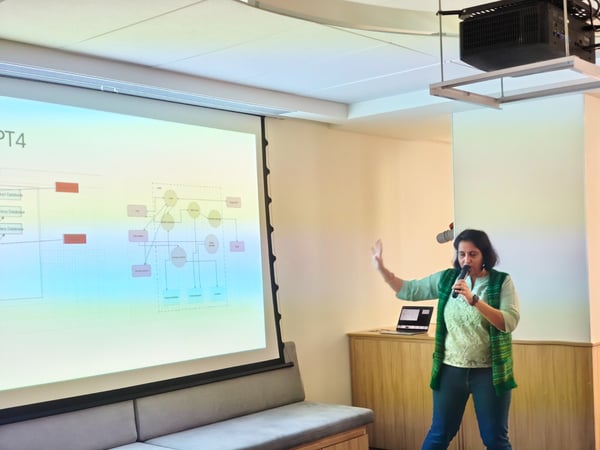 A woman is giving a technical presentation indoors, holding a microphone and gesturing toward a large projection screen that displays a system architecture diagram and the text "PT4".