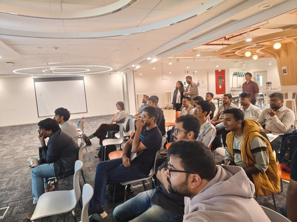 A photograph captures a large group of people, mostly young adults, seated in a modern, brightly lit office space attending a presentation or lecture. The audience is facing a large, blank projection screen mounted on a white wall.
