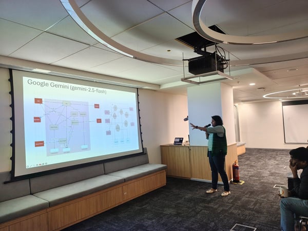 A presenter stands in a modern office or conference room, holding a microphone and gesturing toward a large projection screen displaying a technical flow chart titled "Google Gemini (gemini-2.5-flash)." Several audience members are visible seated on the right side.
