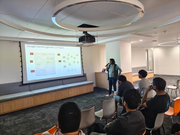 A photograph capturing an indoor presentation in a modern office space, where a presenter stands beside a large screen displaying a technical diagram titled "Google Gemini (gemini-2.5-flash)" to a seated audience.