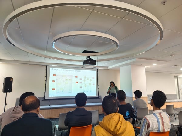 A woman is giving a technical presentation about Google Gemini to an audience seated in a modern, brightly lit conference room, with the presentation slides projected onto a large screen.