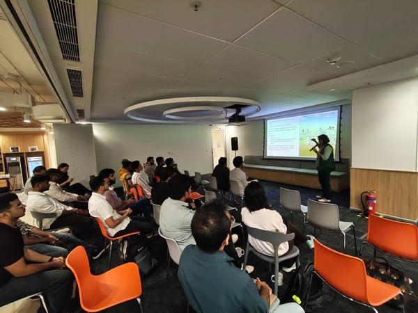 A photograph captures an indoor presentation setting where a speaker stands in front of a projection screen, addressing a large seated audience in a modern office or event space.