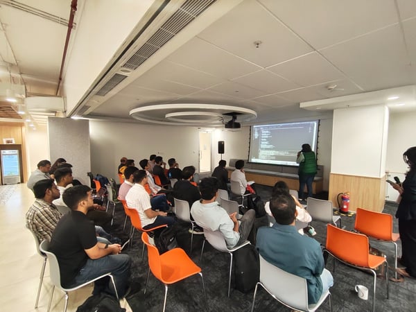 A wide-angle photograph captures a technical presentation or training session taking place in a modern office environment, where a large audience is seated, watching code displayed on a projection screen while an instructor stands nearby.