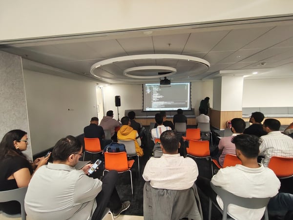 A candid photograph captures an audience seated in a modern indoor conference room, attending a technical presentation where lines of code are displayed on a large projection screen.