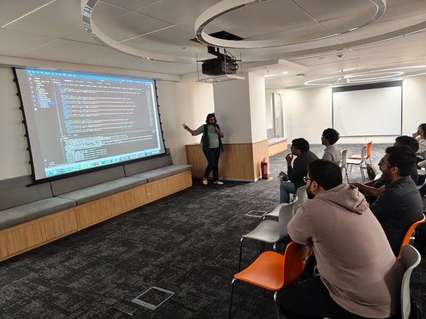 A photograph captures a technical presentation taking place in a modern office meeting room, where a woman stands beside a large screen displaying lines of code to a seated audience.