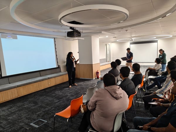A photograph capturing an indoor corporate presentation or meeting, showing a woman speaking into a microphone at the front of the room while a large audience sits facing the screen.