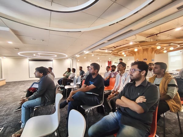 A candid photograph capturing an audience of approximately two dozen people, mostly men, seated in a modern office conference room attending a presentation. The attendees are facing toward a blank projection screen visible on the far left wall.