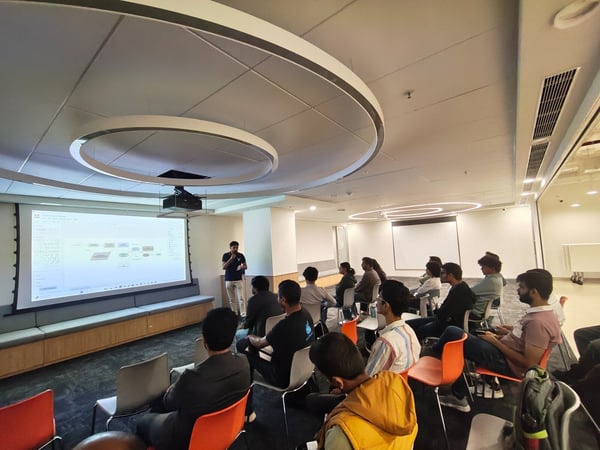 A photograph capturing an indoor corporate presentation where a man stands beside a large projection screen displaying a technical diagram, addressing an audience seated in a modern conference room.