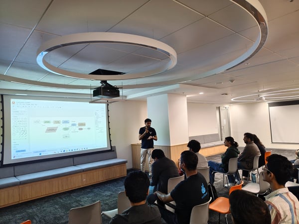 A photograph captures a presentation session taking place in a modern office or seminar room, where a speaker stands beside a large screen displaying a technical diagram to a seated audience.