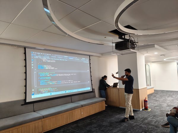 A man is giving a technical presentation using a microphone in a modern office or training room, with lines of code displayed prominently on a large projection screen behind him.