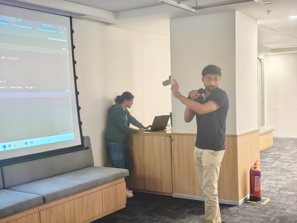 A candid photo captures a technical presentation indoors, featuring a man speaking into a microphone while a woman manages a laptop displaying code on a large projector screen.