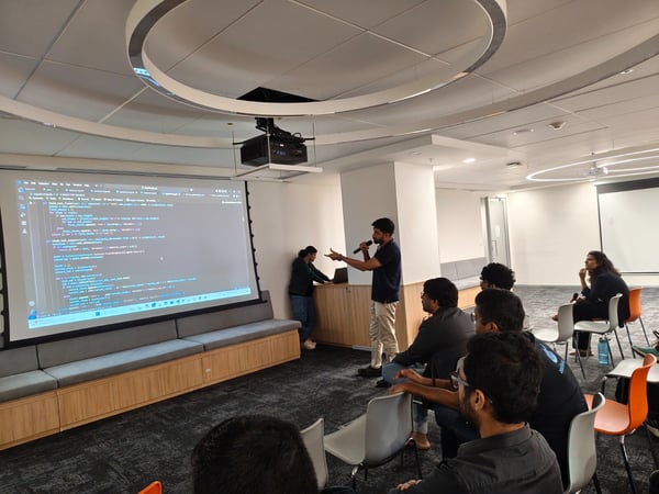 A photograph capturing a technical presentation or workshop in a modern office space, featuring a speaker addressing a seated audience in front of a large projection screen displaying lines of code.
