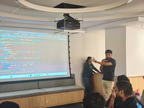 A man is giving a technical presentation, holding a microphone and gesturing toward a large projected screen displaying lines of code, while an audience watches in an indoor setting.