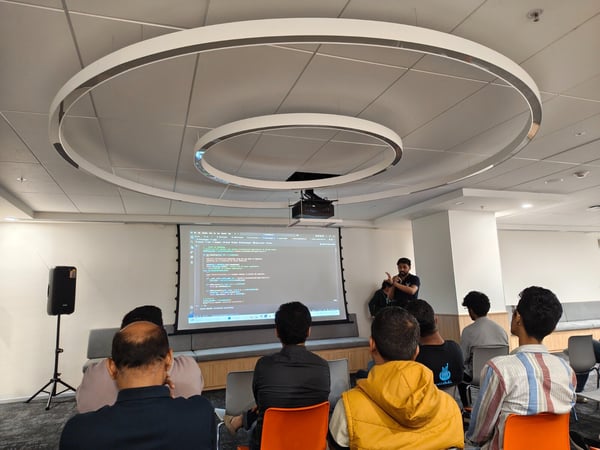 A photograph captures a technical presentation or coding workshop taking place indoors, featuring a presenter standing beside a screen displaying code while an audience sits facing forward.