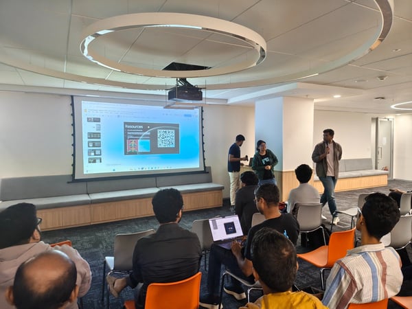A photograph captures an indoor presentation or meeting where a group of people are seated, facing a large projector screen displaying a slide titled "Resources" and a QR code. Three individuals are standing near the screen, appearing to lead the session.