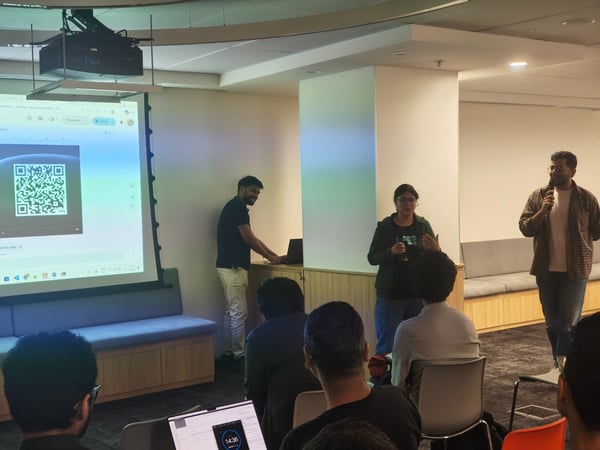 A photograph capturing an indoor technical presentation where a speaker holds a microphone while two other presenters stand nearby, addressing an audience seated in the foreground. A large projector screen displays a QR code and a computer interface.