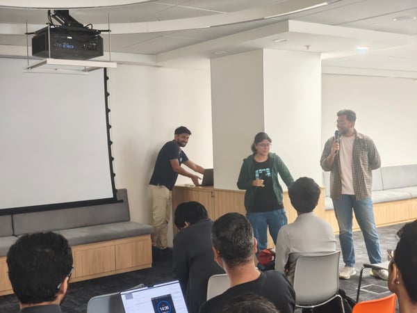 This photograph captures an indoor presentation setting where two speakers, a man and a woman, address an audience in front of a large, blank projection screen, while a third person adjusts equipment nearby.