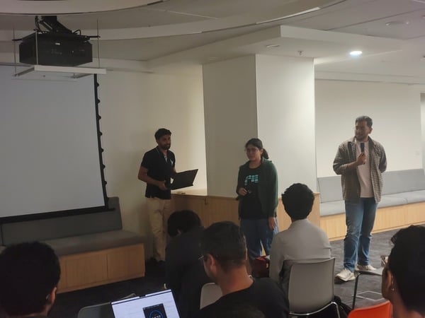 A candid indoor photograph captures a presentation or meeting where three speakers stand near a large blank projection screen, addressing an audience seated in the foreground.