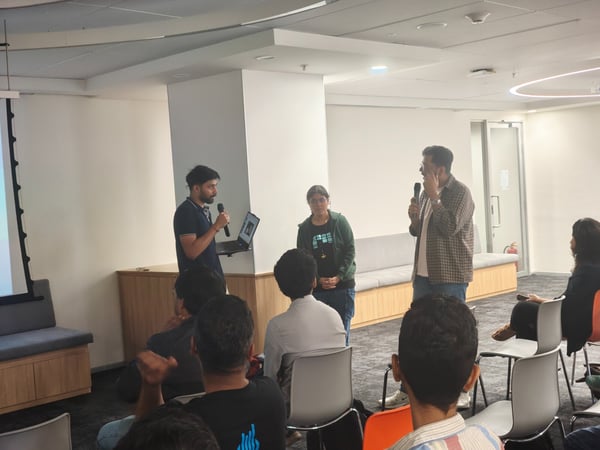 A candid photo capturing an indoor presentation or Q&A session where two men and one woman are standing near a projection screen, speaking into microphones, while an audience is seated in the foreground and mid-ground.