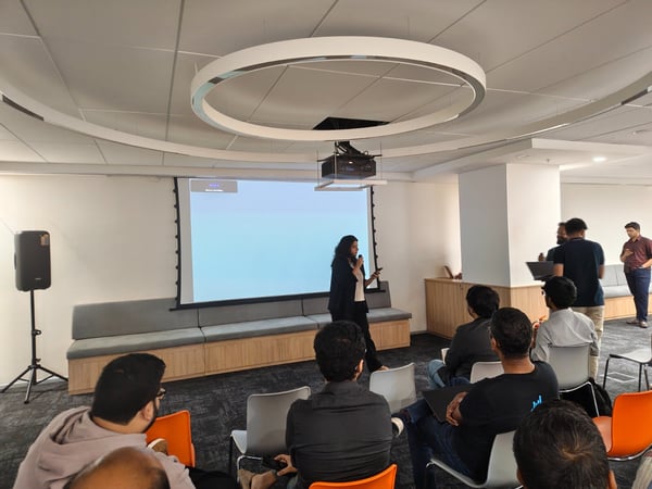 A photograph capturing an indoor corporate presentation where a woman stands next to a large projection screen, addressing a seated audience in a modern office space.