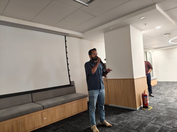 A man wearing glasses and casual attire stands in a modern office space holding a microphone, appearing to give a presentation in front of a large blank projection screen.