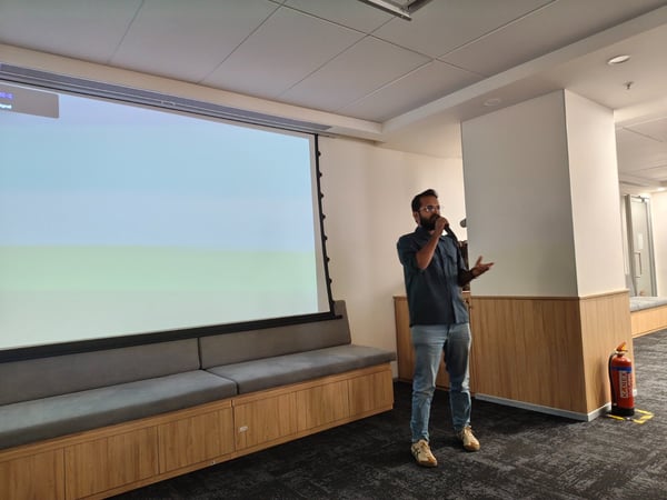 A man wearing glasses and a dark shirt is giving a presentation or talk while holding a microphone in a modern indoor meeting space.