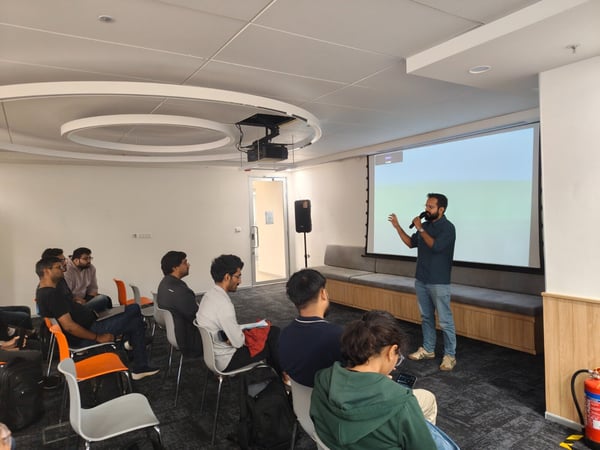 A photograph capturing a presentation or meeting indoors, where a bearded man is speaking into a microphone in front of a blank projection screen while an audience is seated, listening.