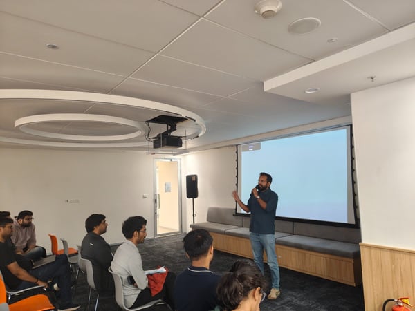 This is a candid photo of a man giving a presentation or talk using a microphone in a modern meeting room, addressing a small audience seated in front of him. The projection screen behind the speaker is illuminated but mostly blank.