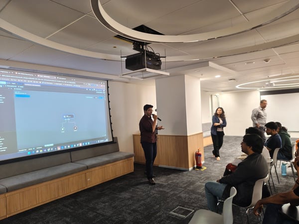 A man is giving a presentation using a microphone in a modern office meeting room, standing next to a large projected screen displaying a software interface, while several attendees are seated in the foreground and background.
