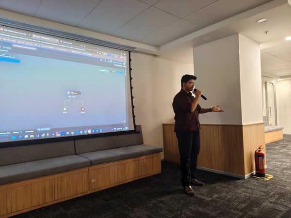 A man wearing a dark shirt and glasses is giving a presentation indoors, holding a microphone and gesturing toward a large projection screen displaying a technical diagram or web application interface.