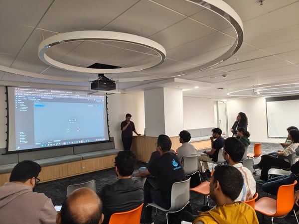 A photograph captures a technical presentation in a modern office or conference room, showing a male presenter standing next to a large projector screen displaying code or a flow diagram while an audience of about 18 people watches.