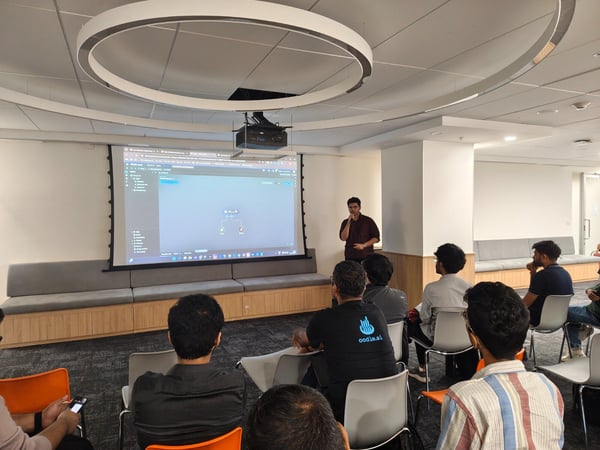 A photograph capturing a technical presentation taking place indoors, where a presenter stands beside a large screen displaying code or a flow diagram to an audience seated in the foreground.